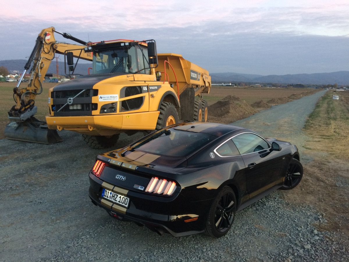 Foto des Rent-A-Racer (schwarzer Sportwagen mit goldenen Zierstreifen) auf einer Baustelle neben einem gelben Bagger. Perspektive aus drei Metern Höhe hinter dem Rent-A-Racer. Geschossen von Joachim Verheugen in 2018.