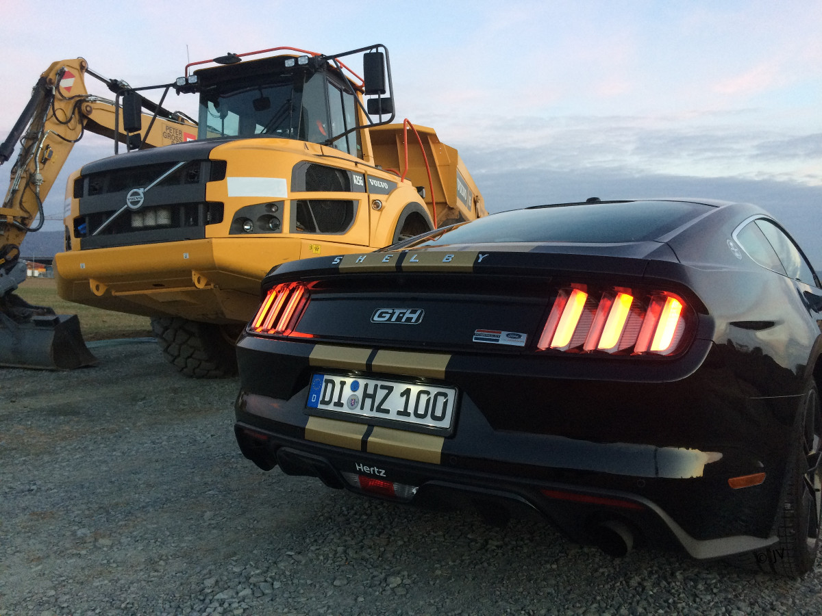 Foto des Rent-A-Racer (schwarzer Sportwagen mit goldenen Zierstreifen) auf einer Baustelle neben einem gelben Bagger. Perspektive hinter dem Rent-A-Racer auf einem halbem Meter Höhe. Geschossen von Joachim Verheugen in 2018.